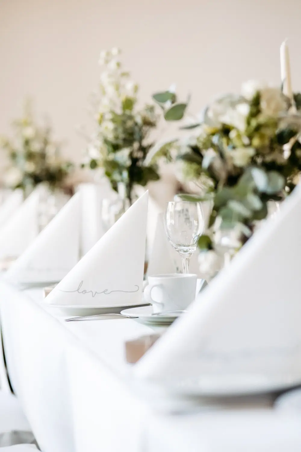 Nahaufnahme eines elegant gedeckten Tisches mit weißen Servietten, die jeweils zu einem Dreieck gefaltet sind, weißen Teetassen, Weingläsern und floralen Tafelaufsätzen mit weißen und grünen Blumen. Die Szene ist hell und sanft fokussiert.