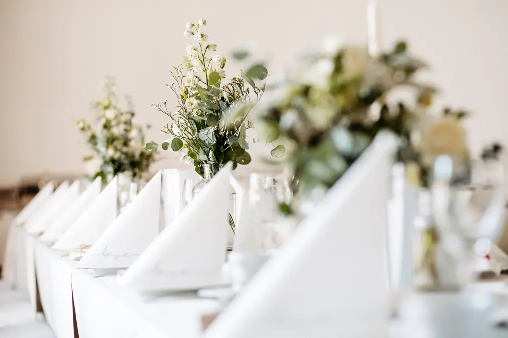 Eine Nahaufnahme eines wunderschön gedeckten Banketttisches mit weißen, zu Dreiecken gefalteten Servietten und eleganten Blumensträußen in Glasvasen, die eine helle und elegante Atmosphäre schaffen.