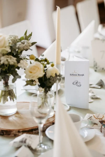 Elegant gedeckter Hochzeitstisch mit weiĂźen Rosen und GrĂĽnpflanzen in Glasvasen, weiĂźen Kerzen, einer MenĂĽkarte mit der Aufschrift Schloss Burgk und ordentlich gefalteten Servietten auf einer hellen Tischdecke.
