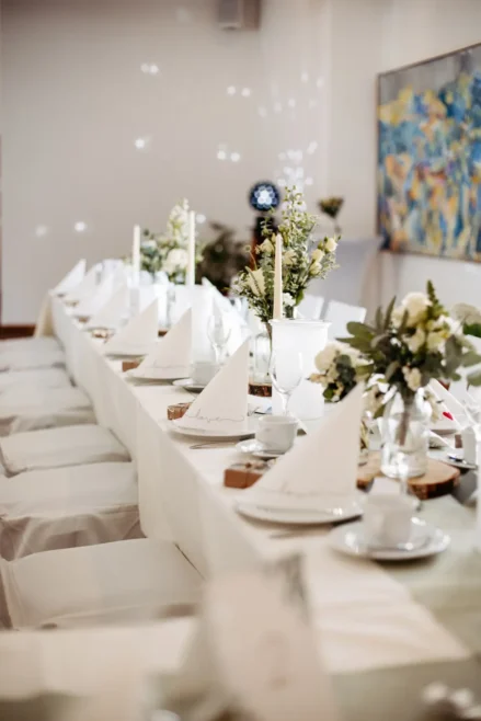 Langer, für eine Veranstaltung gedeckter Banketttisch mit weißer Tischdecke, gefalteten Servietten, Kerzen und floralen Tafelaufsätzen in einem hellen, eleganten Raum mit moderner Kunst an der Wand.