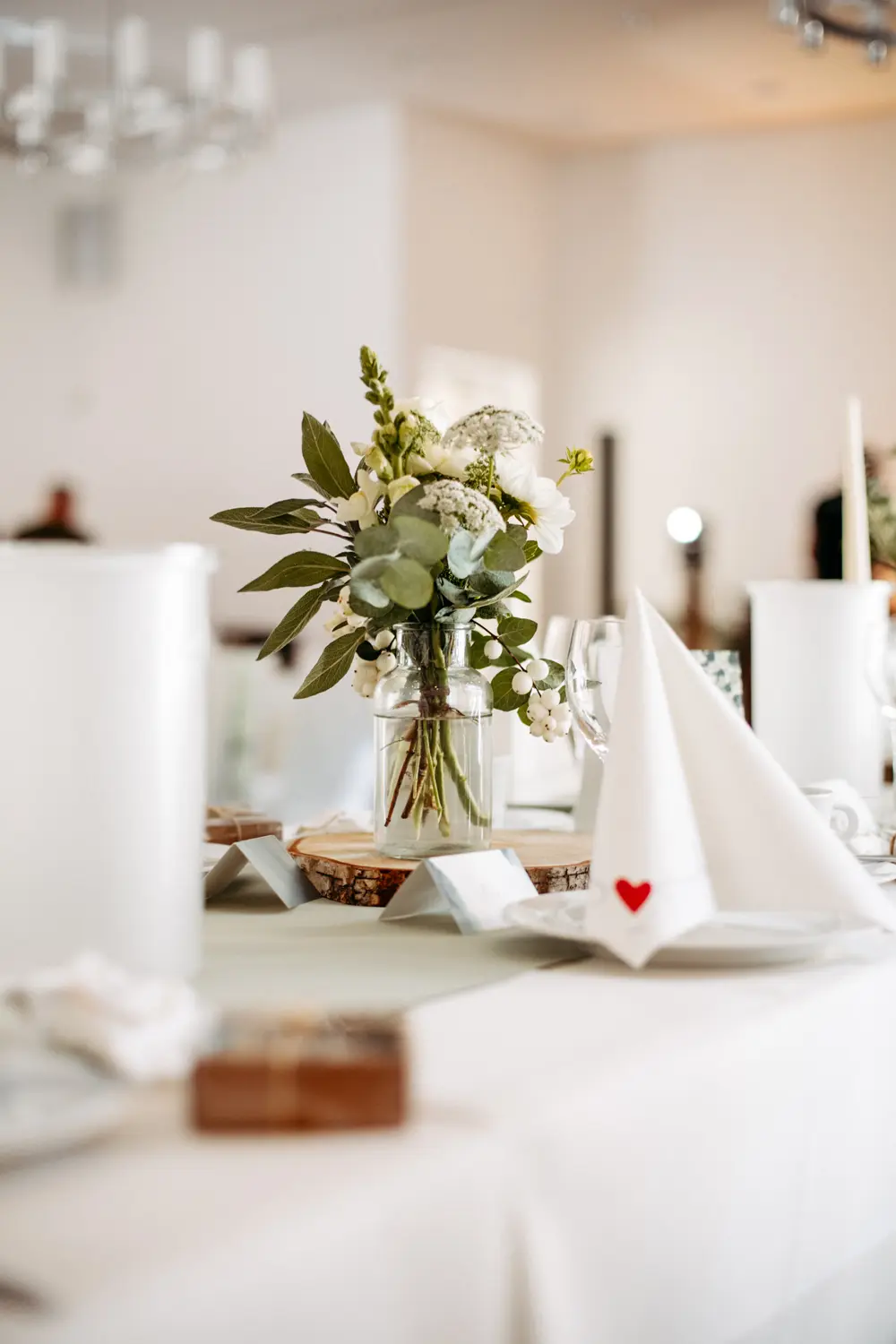 Eine Nahaufnahme eines elegant gedeckten Tisches mit einer Vase mit weiĂźen und grĂĽnen Blumen, gefalteten Servietten mit kleinen roten Herzen und sanfter Beleuchtung in einem hellen, luftigen Raum.