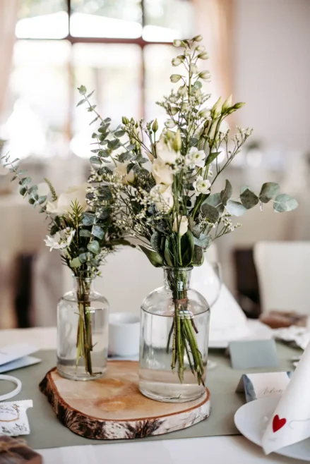 Zwei Glasvasen mit weiĂźen und grĂĽnen Blumen und Eukalyptuszweigen stehen auf einer Holzplatte als Tafelaufsatz auf einem Tisch, mit weichem natĂĽrlichem Licht und unscharfem Hintergrund.