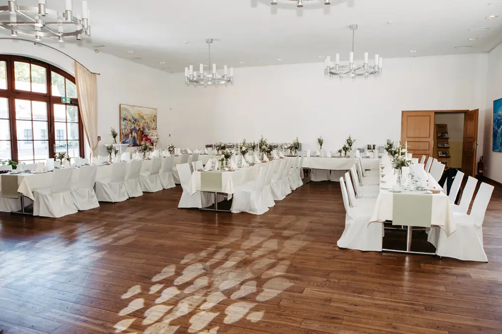 Ein heller Bankettsaal mit Holzböden, weißen Wänden und Kronleuchtern. Drei lange Tische sind mit weißer Bettwäsche und einfachen Blumensträußen eingedeckt, um die herum ordentlich weiß bezogene Stühle stehen. Große Fenster lassen viel natürliches Licht herein.