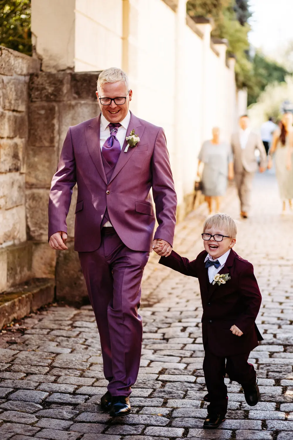Ein Mann und ein Junge, beide in violetten Anzügen und mit Brillen, gehen Hand in Hand und lächeln, während sie einen Kopfsteinpflasterweg entlang schlendern, mit verschwommenen Menschen und Grün im Hintergrund.
