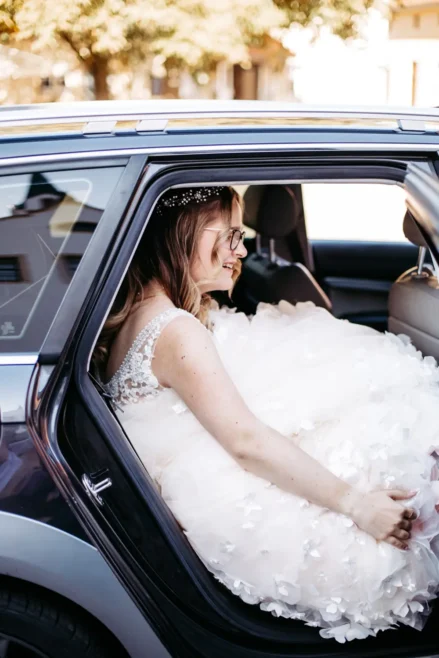Eine Braut in einem weißen Hochzeitskleid mit Blumendetails und einem mit Juwelen besetzten Stirnband sitzt lächelnd auf dem Rücksitz eines Autos mit offener Tür, während das Sonnenlicht auf sie scheint.