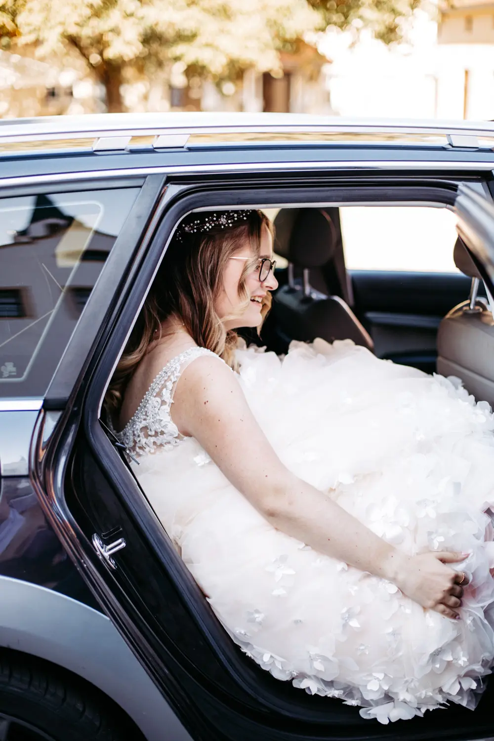 Eine Braut in einem weißen Hochzeitskleid mit Blumendetails und einem mit Juwelen besetzten Stirnband sitzt lächelnd auf dem Rücksitz eines Autos mit offener Tür, während das Sonnenlicht auf sie scheint.