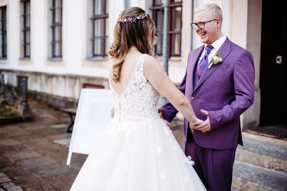 Eine Braut in einem weißen Kleid und ein Bräutigam in einem lila Anzug lächeln sich freudig an, während sie sich vor einem Gebäude mit großen Fenstern an den Händen halten.