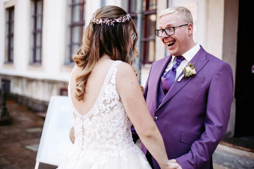 Eine Braut in einem weißen Spitzenkleid hält die Hand eines lächelnden Bräutigams in einem lila Anzug mit Krawatte. Sie stehen sich draußen gegenüber, der Bräutigam sieht überrascht und glücklich aus.