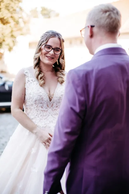 Eine Braut in einem weißen Spitzenhochzeitskleid und mit Brille lächelt einen Mann in einem lila Anzug an, der ihr an einem sonnigen Tag im Freien gegenübersitzt.
