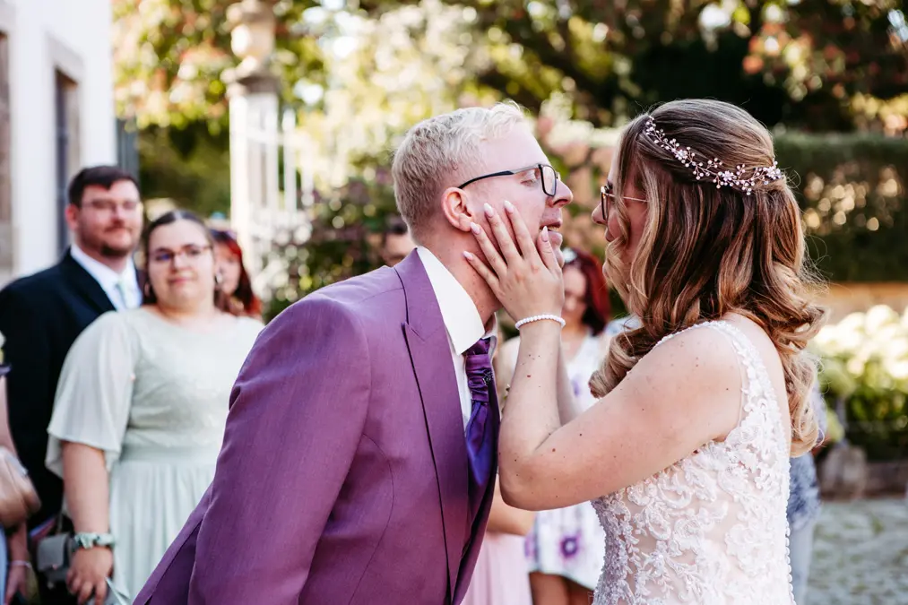 Eine Braut in einem weißen Kleid hält sanft das Gesicht eines Bräutigams in einem lavendelfarbenen Anzug, während sie sich im Freien anlächeln, während die Gäste im Hintergrund zusehen.