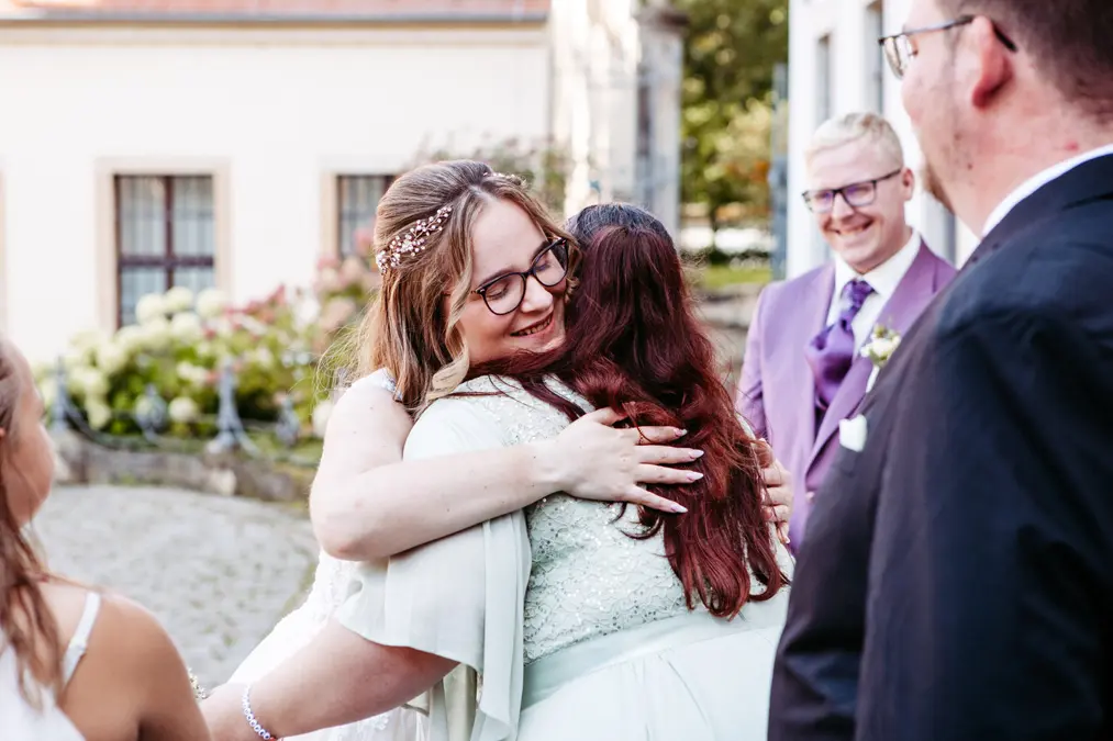 Eine Braut mit Brille und einem schmuckvollen Stirnband umarmt eine Frau mit langen dunklen Haaren. Sie lächeln im Freien, umgeben von formell gekleideten Hochzeitsgästen, darunter ein Mann in einem hellvioletten Anzug.