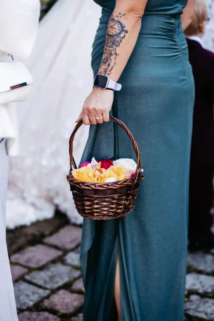 Eine Frau in einem blaugrünen Kleid mit einem tätowierten Arm hält einen mit gelben und rosa Blumen gefüllten Weidenkorb. Sie steht auf einem Steinweg, und im Hintergrund sind teilweise andere Personen zu sehen.