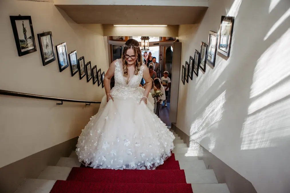 Eine Braut in einem weißen, geblümten Kleid und mit Brille schreitet lächelnd eine Treppe mit rotem Teppich hinauf. Ihr folgen Brautjungfern und Gäste. Gerahmte Fotos säumen die Wände des Treppenhauses. Das Sonnenlicht wirft Schatten an die Wand.