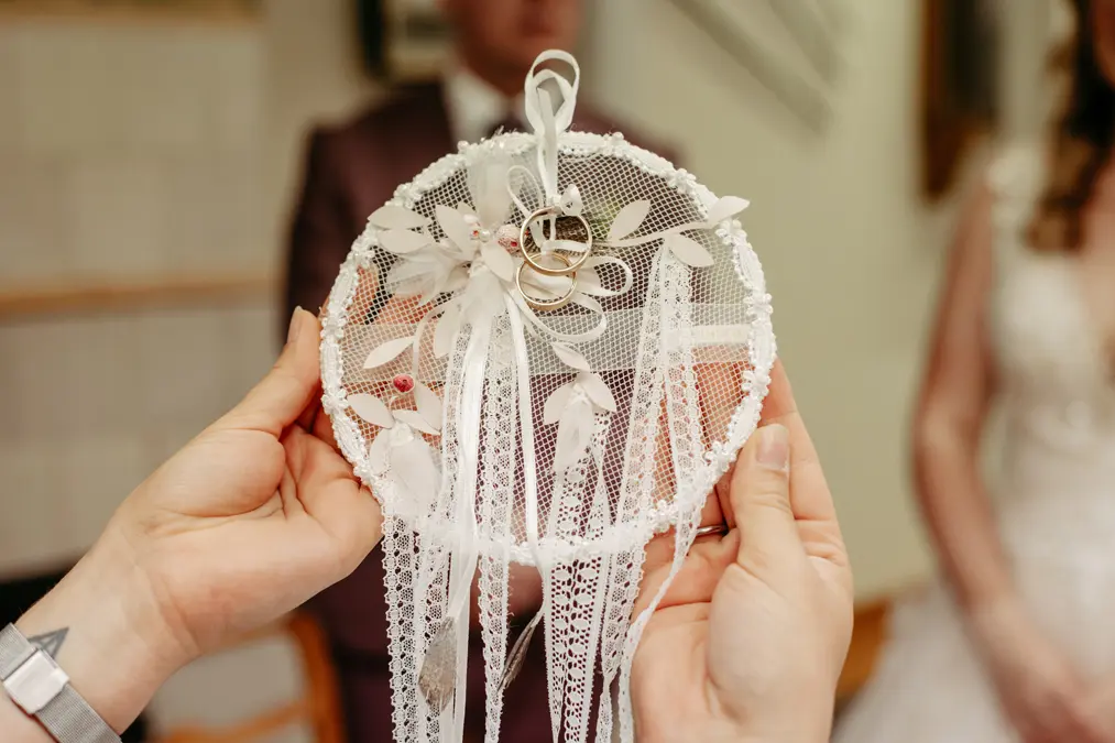 Eine Person hält einen mit Blumen und Bändern geschmückten Traumfänger aus Spitze, auf dem zwei goldene Eheringe zu sehen sind, während Braut und Bräutigam im Hintergrund verschwommen zu sehen sind.