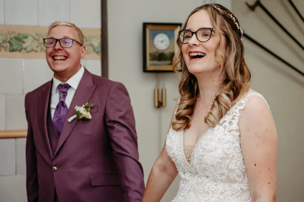 Eine lächelnde Braut im weißen Spitzenkleid und mit Brille steht neben einem Mann in lila Anzug und Krawatte, beide wirken glücklich und fröhlich im Haus.
