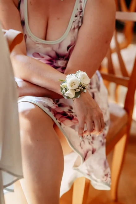 Eine Frau in einem ärmellosen, geblümten Kleid sitzt mit verschränkten Armen da und zeigt eine weiße Rosenkorsage an ihrem Handgelenk. Sie sitzt auf einem Holzstuhl in einem warm beleuchteten Raum.