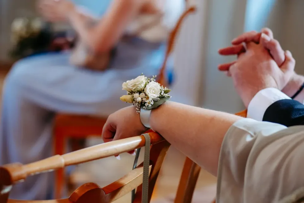 Eine Person, die ein weißes Rosenkorsett am Handgelenk trägt, stützt ihren Arm auf einen Holzstuhl, während eine andere Person unscharf im Hintergrund sitzt.