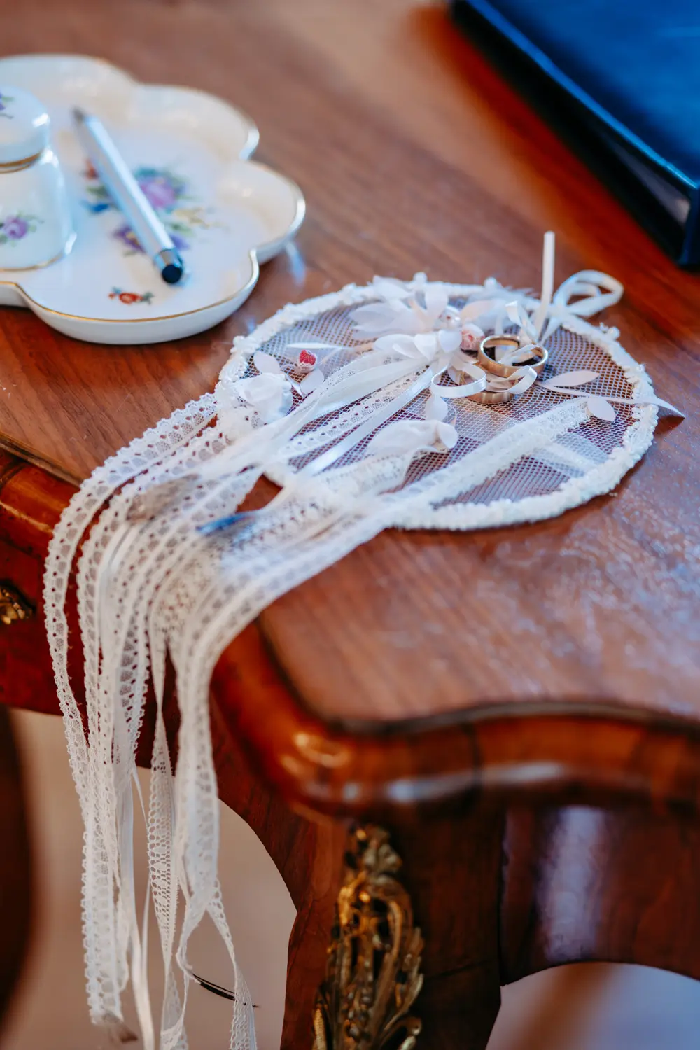 Ein zarter Traumfänger aus weißer Spitze mit Bändern und zwei Eheringen liegt auf einem polierten Holztisch neben einer geblümten Porzellanschale und einem Stift.