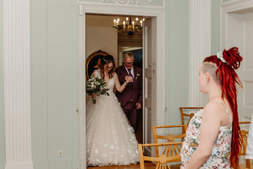 Eine Braut in einem weißen Kleid und ein Mann im Anzug gehen Arm in Arm durch eine Tür, während eine Frau mit roten Haaren in einem geblümten Kleid in einem geschmückten Raum von der Seite zusieht.