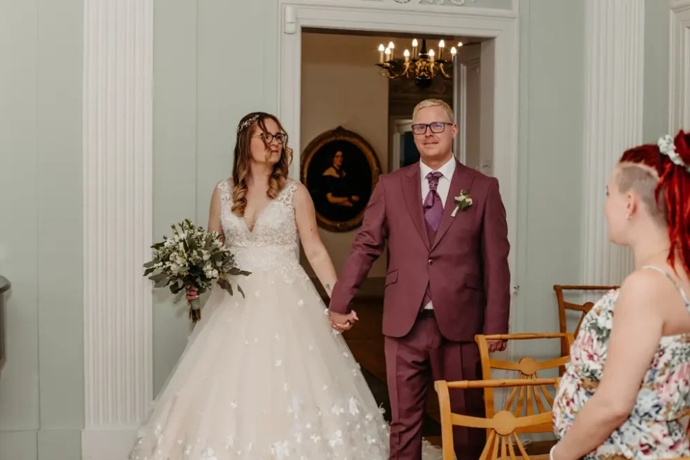 Eine Braut in einem weißen Kleid mit einem Blumenstrauß in der Hand und ein Bräutigam in einem lilafarbenen Anzug halten sich an den Händen, während ein rothaariger Gast in einem Innenraum neben leeren Stühlen steht. Hinter den beiden hängt ein Porträt an der Wand.