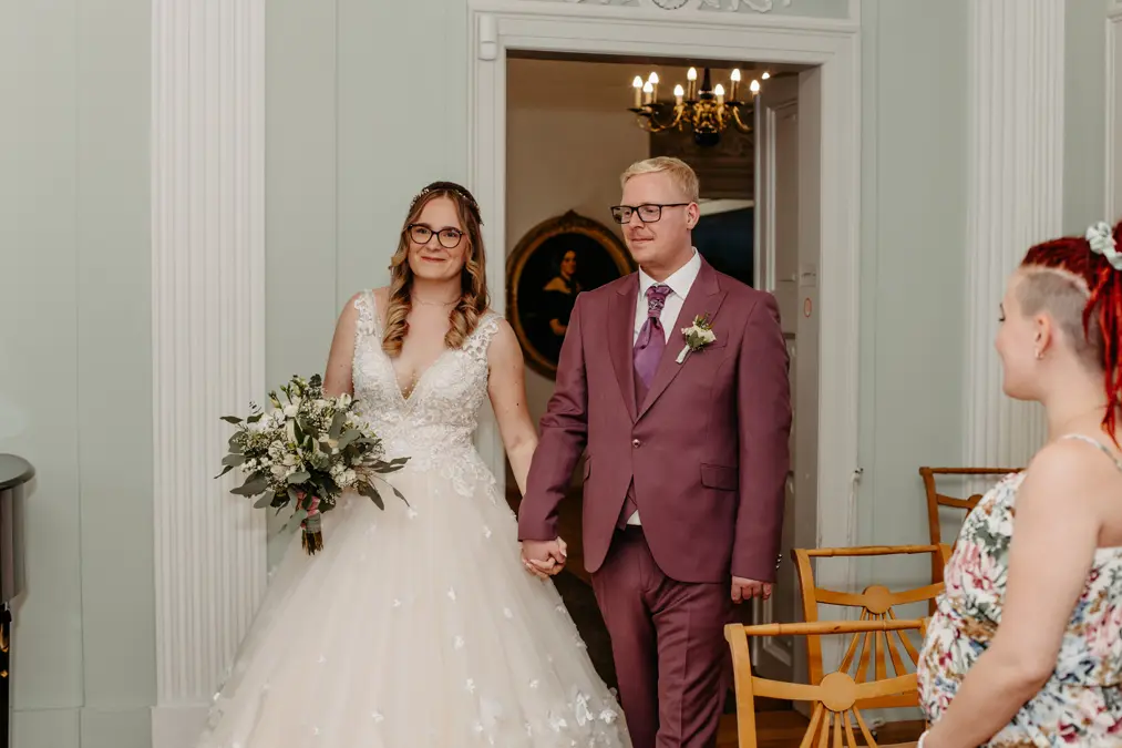 Eine Braut in einem weißen Kleid, die einen Blumenstrauß hält, und ein Bräutigam in einem lilafarbenen Anzug halten sich an den Händen und stehen zusammen in einem Innenraum. Eine Frau mit roten Haaren, von der Seite gesehen, sitzt in der Nähe und beobachtet die beiden.