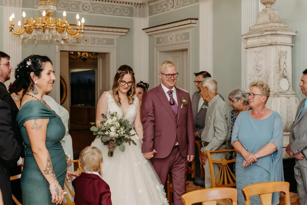 Eine Braut in einem weißen Kleid und ein Bräutigam in einem lilafarbenen Anzug schreiten gemeinsam lächelnd den Gang hinunter, umgeben von stehenden Gästen in formeller Kleidung in einem eleganten Raum mit kunstvollem Dekor.