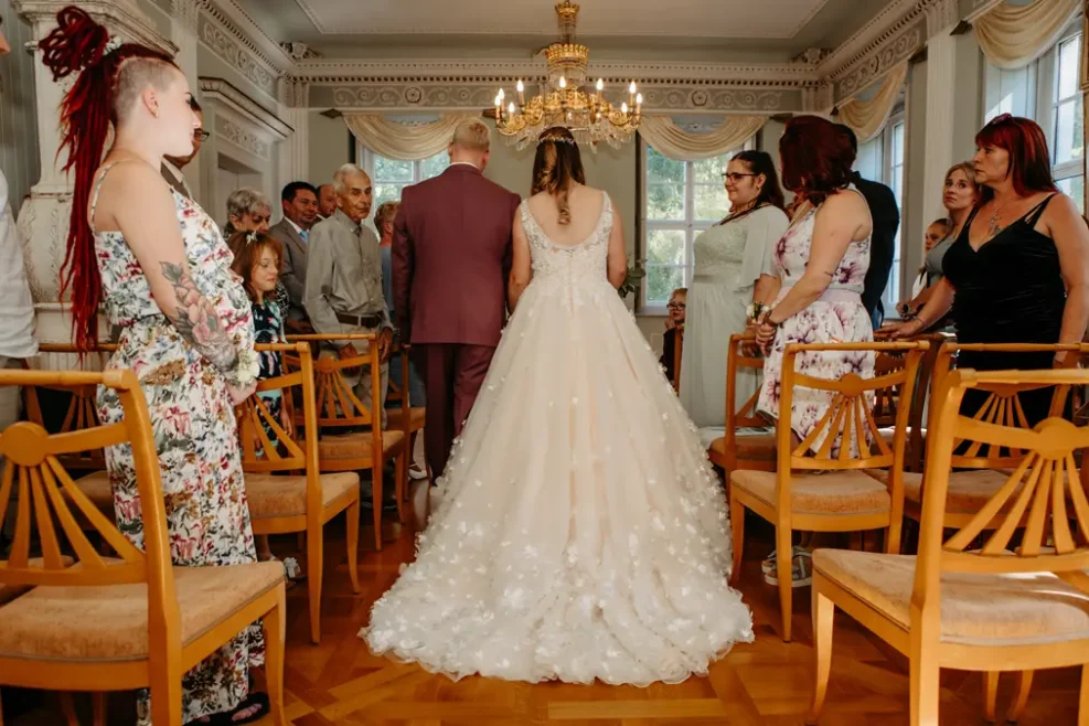 Eine Braut in einem weißen Kleid schreitet mit einem Mann in einem lilafarbenen Anzug zum Altar, umgeben von sitzenden und stehenden Hochzeitsgästen in einem hellen, eleganten Raum mit Holzstühlen und einem Kronleuchter.