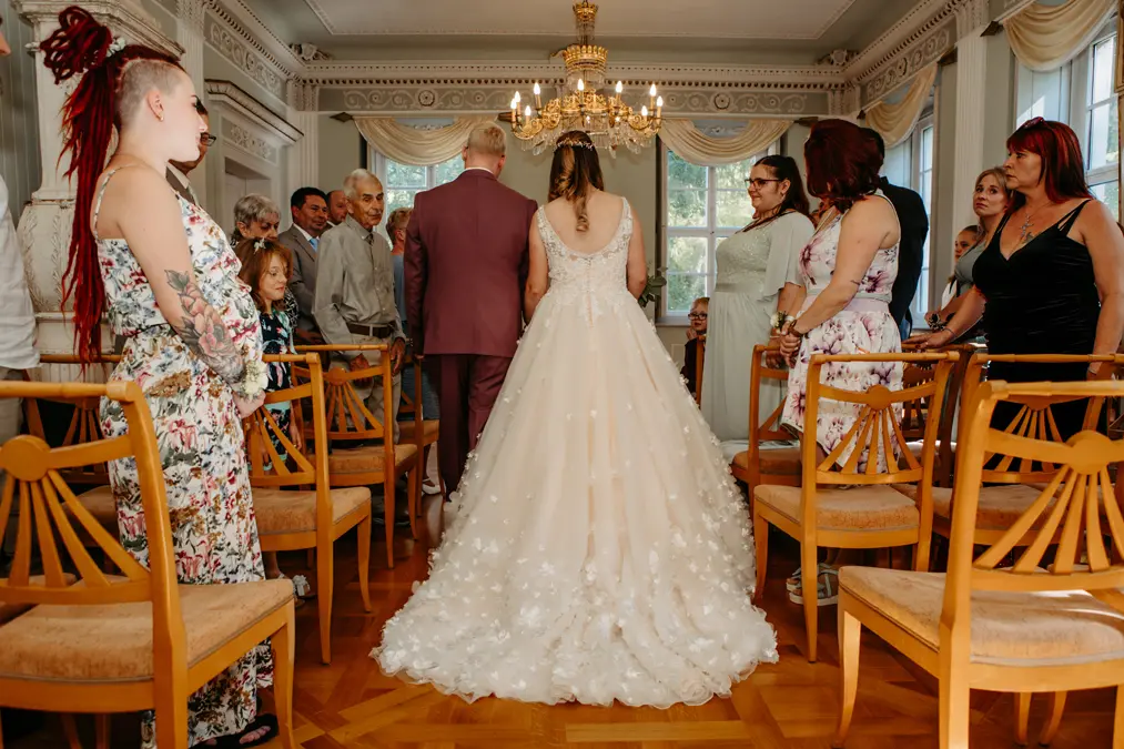 Eine Braut in einem weißen Kleid schreitet mit einem Mann in einem lilafarbenen Anzug zum Altar, umgeben von sitzenden und stehenden Hochzeitsgästen in einem hellen, eleganten Raum mit Holzstühlen und einem Kronleuchter.