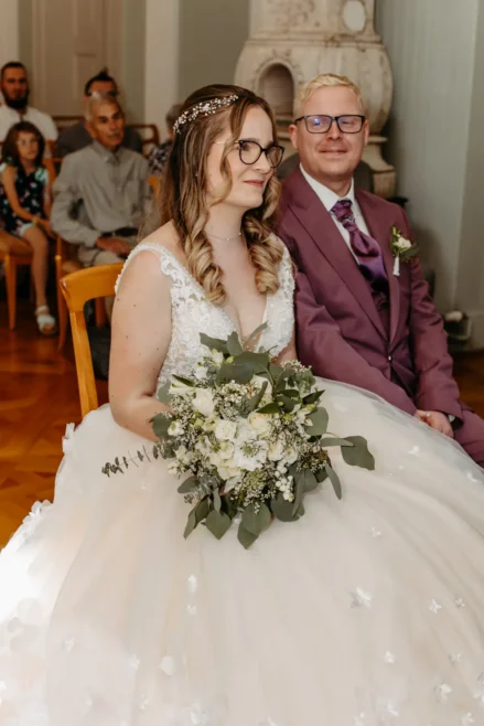 Eine Braut in einem weißen Kleid mit einem Strauß weißer Blumen sitzt neben einem Bräutigam in einem violetten Anzug; sie befinden sich in einem Innenraum, die Gäste sitzen hinter ihnen.