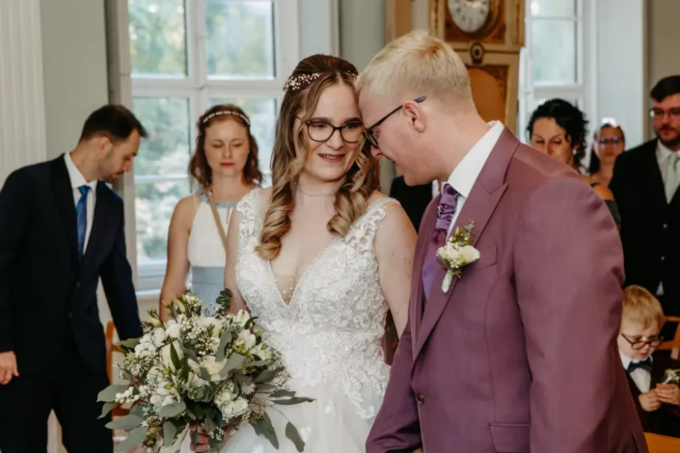 Eine Braut in einem weißen Kleid mit einem Blumenstrauß in der Hand lächelt den Bräutigam in einem lilafarbenen Anzug und mit Brille während einer Hochzeitszeremonie an, während die Gäste im Hintergrund zusehen.