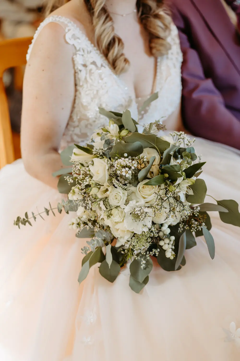 Eine Braut im spitzenbesetzten Hochzeitskleid hält einen Strauß aus weißen Rosen, Schleierkraut und Grünzeug in der Hand, wobei ihre weichen Locken im Haar zu sehen sind.