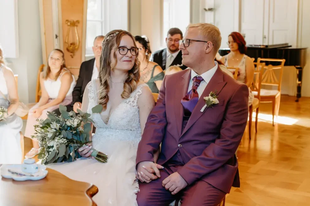 Eine Braut in einem weißen Kleid mit einem Blumenstrauß sitzt Hand in Hand mit einem Bräutigam in einem kastanienbraunen Anzug. Sie lächeln sich während einer Hochzeitszeremonie an, während die Gäste hinter ihnen in einem sonnenbeschienenen Raum Platz nehmen.