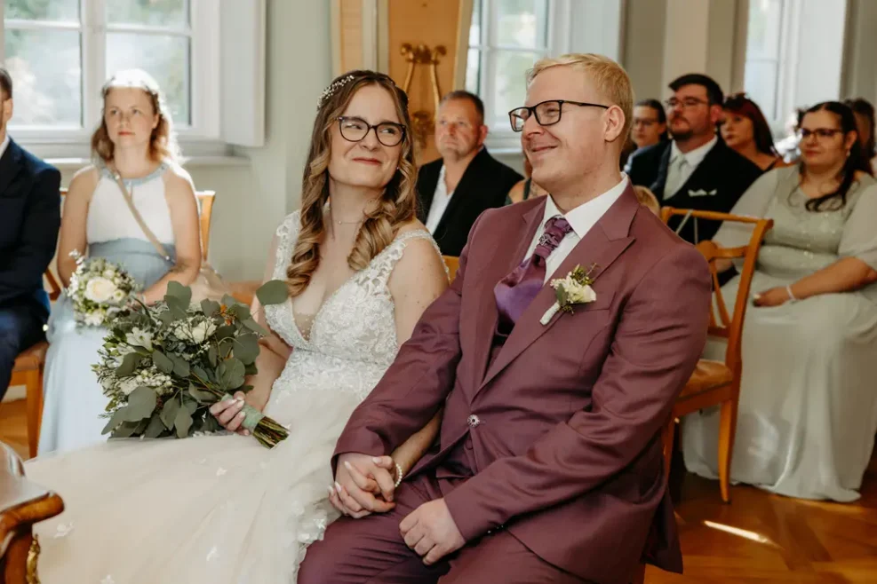Eine Braut in einem weißen Kleid und ein Bräutigam in einem lilafarbenen Anzug sitzen händchenhaltend und lächeln sich während einer Hochzeitszeremonie an, umgeben von sitzenden Gästen in einem hellen Raum.