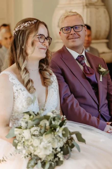 Eine Braut in einem weißen Kleid mit einem Blumenstrauß in der Hand sitzt neben einem Bräutigam in einem lilafarbenen Anzug mit Brille. Beide lächeln und schauen sich während ihrer Hochzeitszeremonie an.