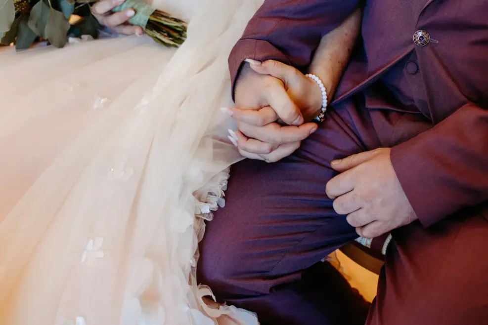 Nahaufnahme eines Paares, das sich an den Händen hält. Die eine trägt ein weißes Hochzeitskleid, die andere einen lilafarbenen Anzug, der ihre Vereinigung an ihrem Hochzeitstag symbolisiert. Das Kleid der Braut hat zarte Blumendetails.