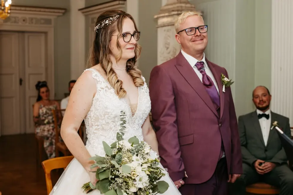 Eine Braut in einem weißen Kleid, die einen Blumenstrauß hält, steht neben einem lächelnden Bräutigam in einem lilafarbenen Anzug. Sie scheinen bei einer Hochzeitszeremonie im Haus zu sein, mit Gästen, die im Hintergrund sitzen.