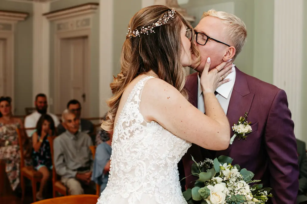 Eine Braut und ein Bräutigam küssen sich während ihrer Hochzeitszeremonie. Die Braut trägt ein Spitzenkleid und ein geblümtes Haarteil, während der Bräutigam eine Brille und einen kastanienbraunen Anzug trägt. Die Gäste sitzen im Hintergrund.