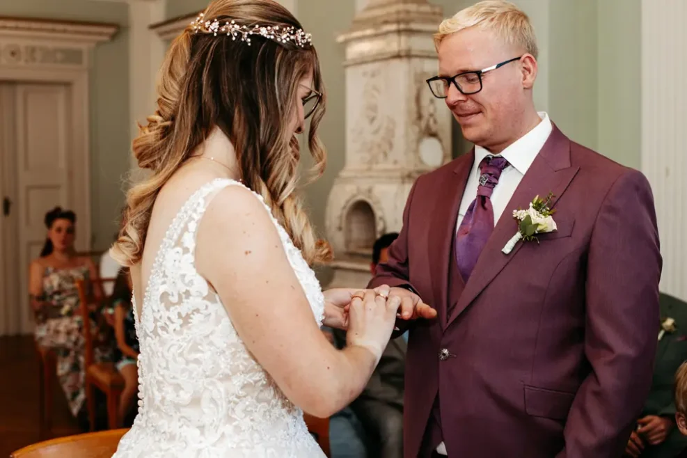 Eine Braut in einem weißen Spitzenkleid steckt dem Bräutigam während einer Hochzeitszeremonie einen Ring an den Finger. Der Bräutigam trägt einen malvenfarbenen Anzug und eine Brille. Die Gäste sitzen im Hintergrund.