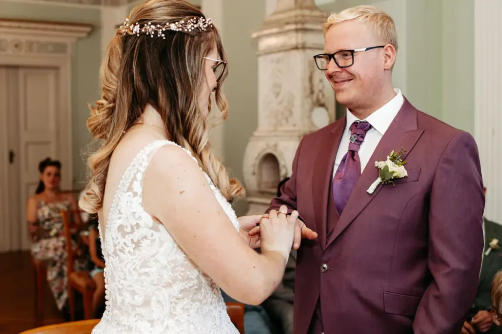 Eine Braut in einem weißen Spitzenkleid steckt dem Bräutigam einen Ring an den Finger. Der Bräutigam trägt einen violetten Anzug mit einer Boutonniere und einer Brille. Sie lächeln sich in einem formellen Innenraum an.