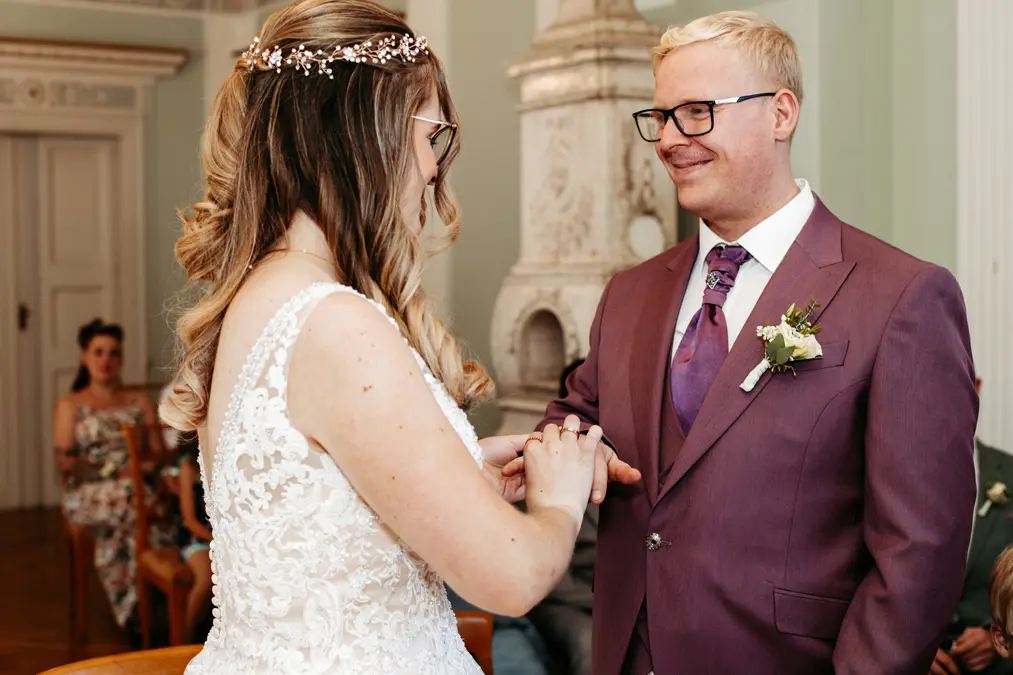Eine Braut in einem weißen Spitzenkleid steckt dem Bräutigam einen Ring an den Finger. Der Bräutigam trägt einen violetten Anzug mit einer Boutonniere und einer Brille. Sie lächeln sich in einem formellen Innenraum an.
