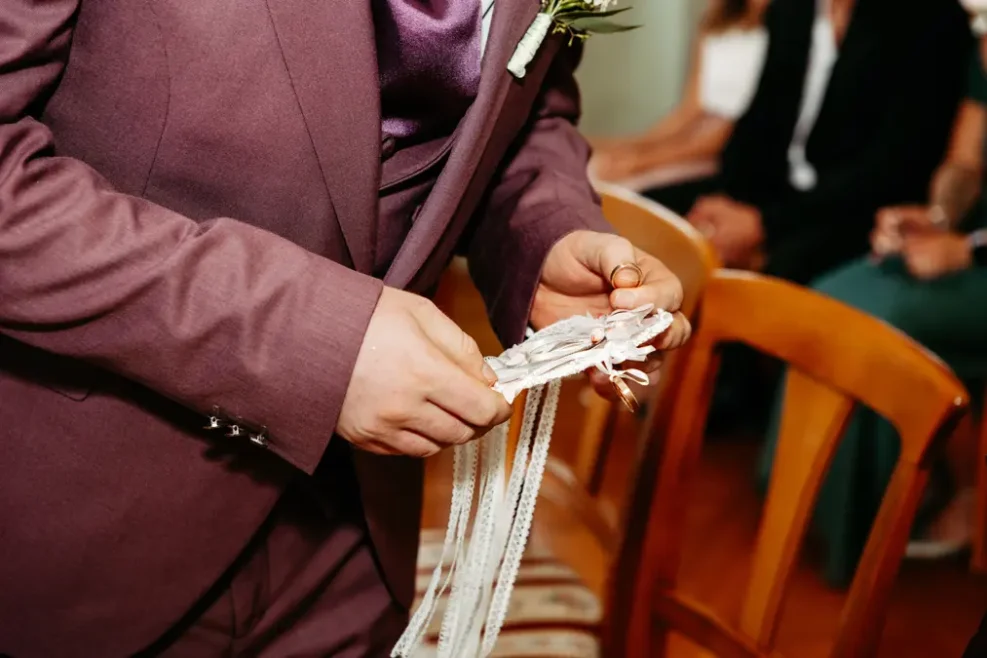 Eine Person in einem lilafarbenen Anzug hält mehrere weiße Schleifenbänder und steht neben Holzstühlen, während andere Personen im Hintergrund sitzen.