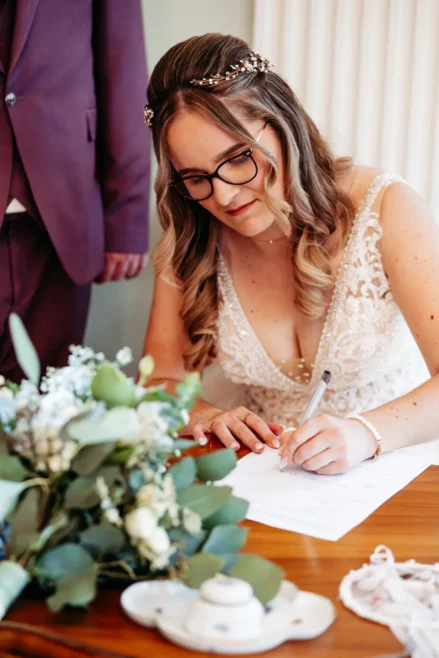 Eine Braut im Spitzenhochzeitskleid und mit Brille unterschreibt ein Dokument an einem mit Blumen geschmückten Tisch. Eine weitere Person in einem Anzug steht in der Nähe, teilweise sichtbar.