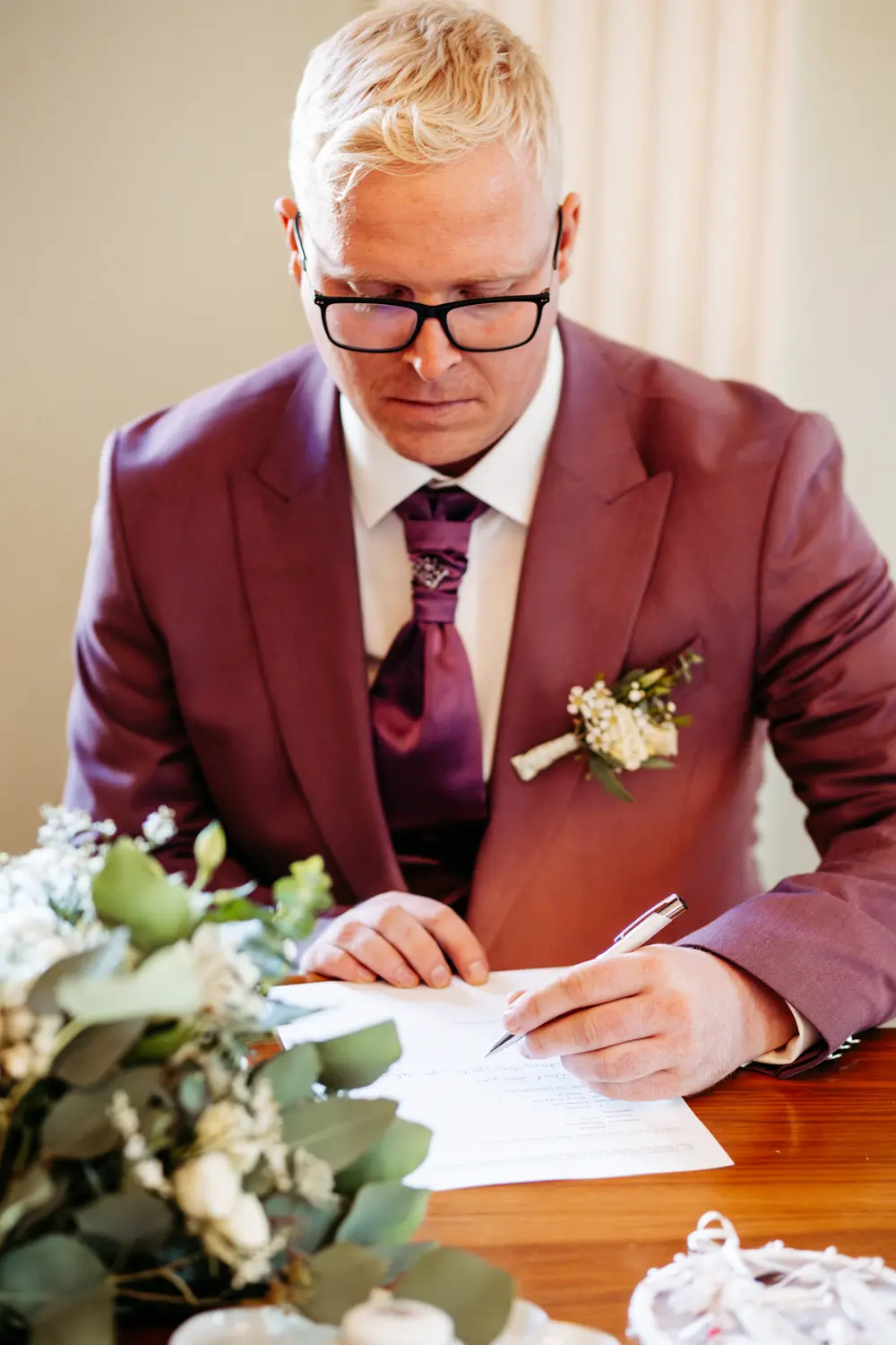 Ein Mann mit blondem Haar und Brille, lilafarbenem Anzug, Krawatte und Ansteckblume sitzt an einem Tisch und unterschreibt ein Dokument, im Vordergrund ein BlumenstrauĂź.