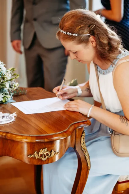 Eine Frau in einem weiĂźen Kleid sitzt an einem verzierten Holztisch und unterschreibt ein Dokument. Auf dem Tisch liegt ein BlumenstrauĂź, und im Hintergrund stehen weitere Personen, die teilweise sichtbar sind.