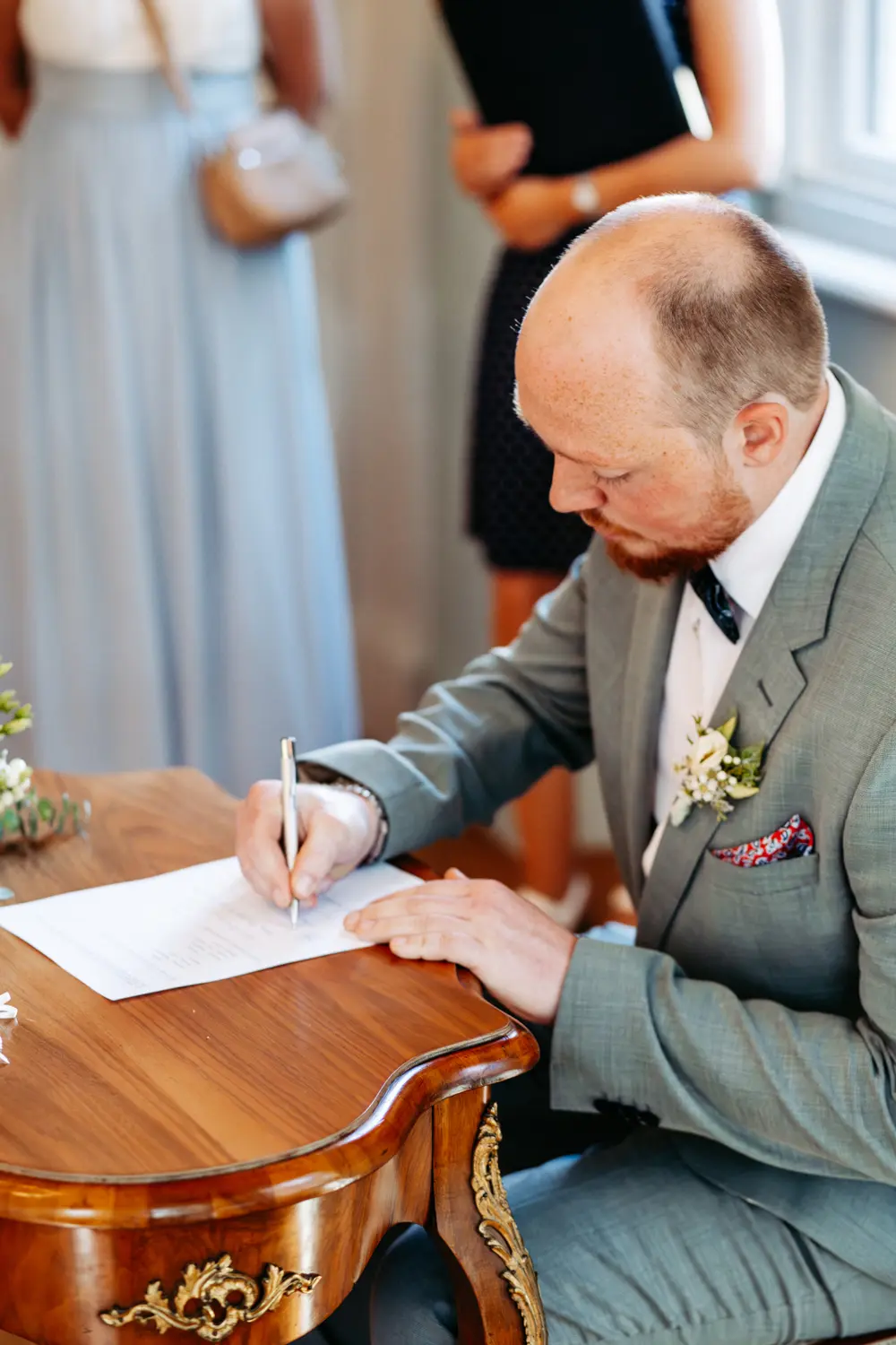 Ein Mann in einem hellgrauen Anzug sitzt an einem Holztisch und unterschreibt ein Dokument mit einem Stift. Er trägt eine Boutonniere an seinem Jackett, was auf eine formelle Veranstaltung oder eine Hochzeit hindeutet. Andere Personen stehen im Hintergrund.