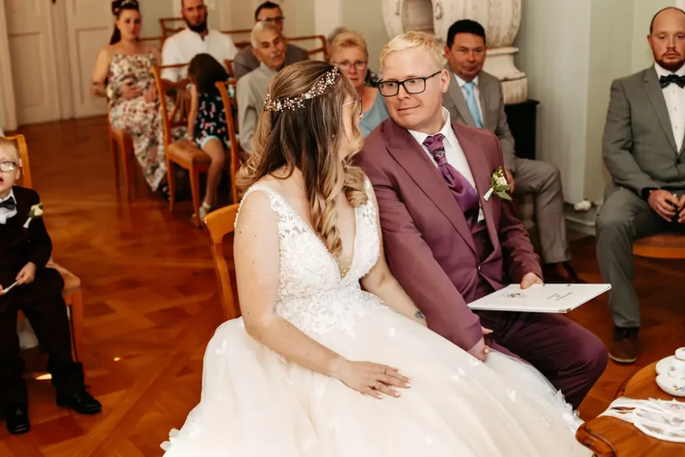Eine Braut und ein Bräutigam sitzen bei ihrer Trauung zusammen und schauen sich an. Die Braut trägt ein weißes Kleid und der Bräutigam einen malvenfarbenen Anzug. Hinter ihnen sitzen Gäste, die in einem geschmückten Raum aufmerksam zusehen.