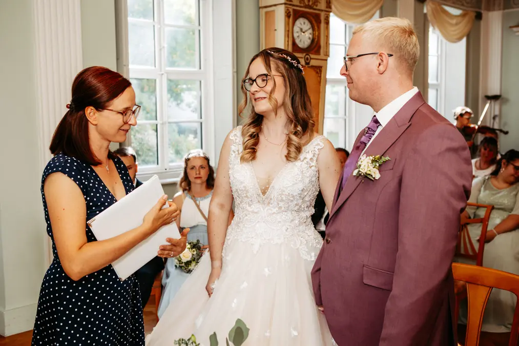 Eine Braut in einem weißen Kleid und ein Bräutigam in einem lilafarbenen Anzug stehen Seite an Seite, lächeln und unterhalten sich mit einer Frau, die in einem hellen Raum mit großen Fenstern und sitzenden Gästen Papiere hält.