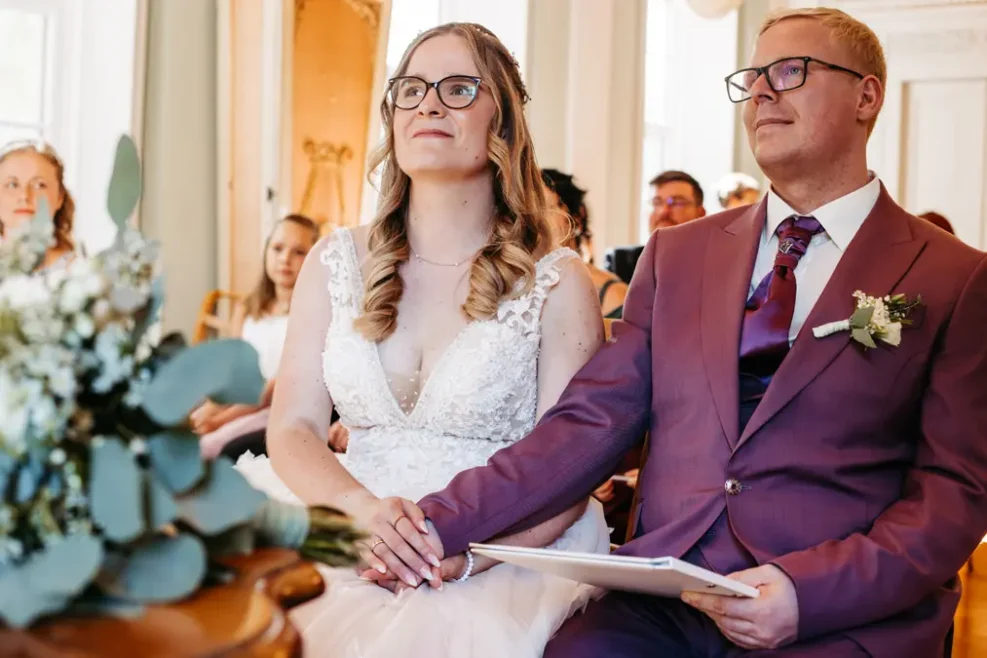 Eine Braut und ein Bräutigam sitzen nebeneinander und halten sich während ihrer Hochzeitszeremonie an den Händen. Die Braut trägt ein weißes Kleid und eine Brille, während der Bräutigam einen lilafarbenen Anzug und eine Brille trägt. Die Gäste sitzen im Hintergrund.