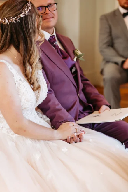 Eine Braut in einem weißen Spitzenkleid und ein Bräutigam in einem lilafarbenen Anzug halten sich während einer Hochzeitszeremonie an den Händen, während im Hintergrund eine weitere Person in einem grauen Anzug sitzt.