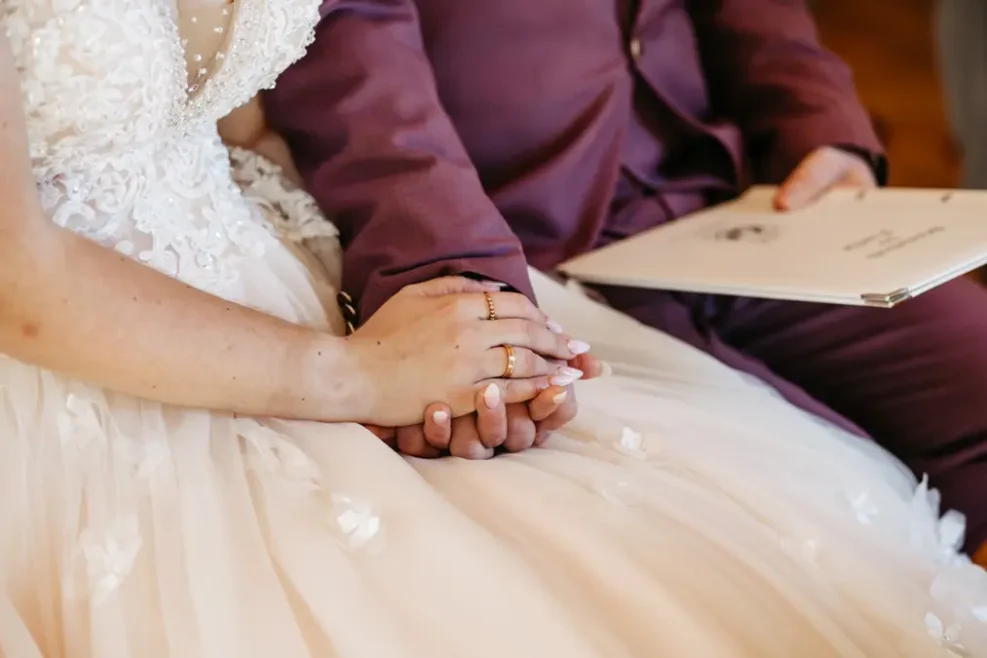 Eine Braut und ein Bräutigam sitzen eng beieinander und halten sich an den Händen. Die Braut trägt ein weißes Spitzenhochzeitskleid, der Bräutigam einen lilafarbenen Anzug und hält eine Mappe oder ein Heft in der Hand. Ihre Eheringe sind sichtbar.